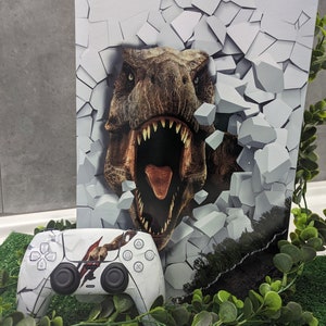 Może przedstawiać: Biała konsola PlayStation 5 z niestandardową skórką przedstawiającą dinozaura przełamującego ścianę. Konsola znajduje się obok białego kontrolera PlayStation 5 z dopasowaną skórką dinozaura.