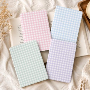 Könnte beinhalten: Vier rechteckige Notizbücher mit einem Vichy-Karo-Muster in Pastellrosa, Hellblau, Mintgrün und Lavendel. Die Notizbücher sind in einem Raster angeordnet und liegen auf einem aufgeschlagenen Buch, dessen gedruckter Text auf den Seiten sichtbar ist.