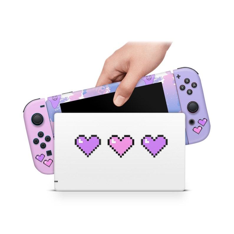 May include: A pink and purple Nintendo Switch console with a pixelated heart design. The console is closed and in a white case with three pixelated hearts on the front.