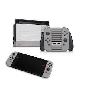 Puede incluir: Una consola Nintendo Switch gris y negra con un diseño personalizado de NES. La consola está cerrada y la pantalla está apagada. Los mandos Joy-Con están conectados a la consola. La consola está sobre una superficie blanca.