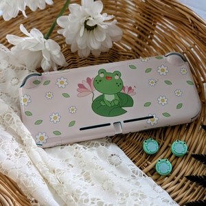May include: A pink Nintendo Switch Lite case featuring a green frog on a lily pad design, surrounded by white daisies and green leaves. Three frog-shaped thumb grips are also visible. The case and grips are set in a wicker basket with white flowers and lace.