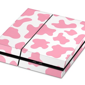 Könnte beinhalten: Eine Spielkonsole mit einem weißen und rosa Kuhmuster. Die Konsole ist rechteckig mit geteilter Oberseite und sichtbaren USB-Anschlüssen. Die rosa Flecken sind unregelmäßig geformt und über die weiße Oberfläche verstreut.