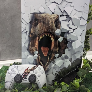 Può includere: Una console di gioco e un controller wireless abbinato con una stampa grafica di un dinosauro T-Rex che rompe un muro bianco incrinato. Il design mostra il dinosauro con la bocca aperta e denti affilati su una superficie frantumata.