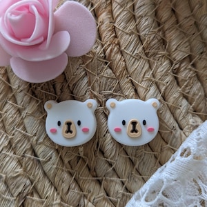 May include: Two white bear-shaped resin buttons with brown noses and pink cheeks. The buttons are on a woven straw background.