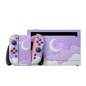 Op de afbeelding: Een lavendelkleurige gameconsole met bijpassende controllers. De console en controllers hebben een ontwerp van witte wolken en halve manen. De controllers hebben zwarte en blauwe knoppen. Het scherm is zwart.