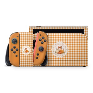 Könnte beinhalten: Eine Spielkonsole und zwei Controller mit einem hellorangen und weißen Vichy-Muster. Die Oberfläche ist mit einem kreisförmigen, weißen spitzenartigen Emblem verziert, das ein Kürbis-Arrangement und eine kleine braune Schleife zeigt.