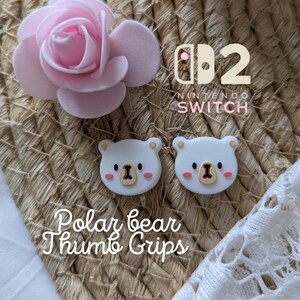 May include: Two white polar bear thumb grips for a Nintendo Switch controller, with a light brown nose and pink cheeks. The image also includes a pink rose and the text "Polar bear Thumb Grips".