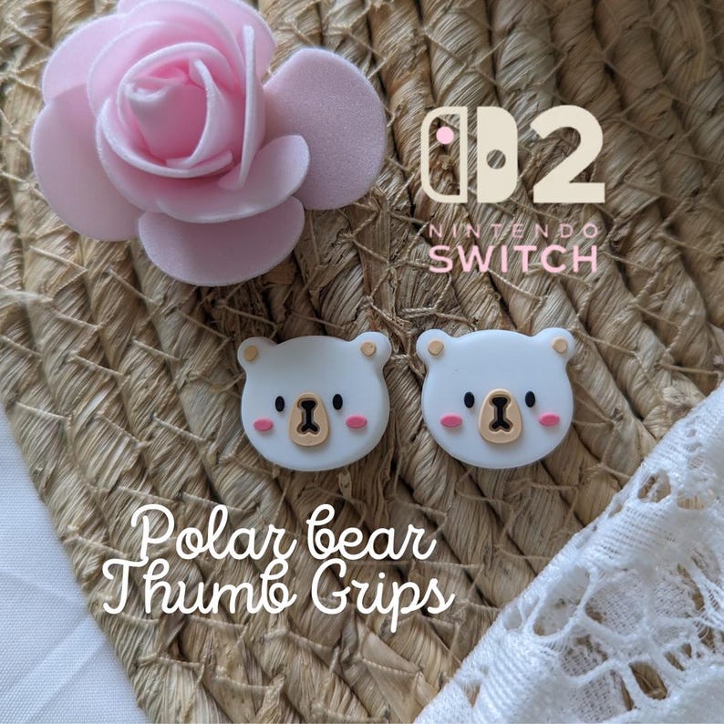 May include: Two white polar bear thumb grips for a Nintendo Switch controller, with a light brown nose and pink cheeks. The image also includes a pink rose and the text "Polar bear Thumb Grips".
