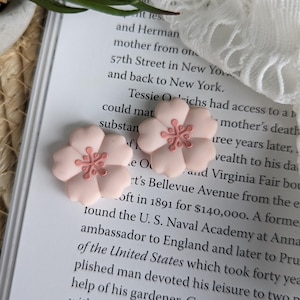 May include: A pair of pale pink flower earrings with a darker pink centre, displayed on an open book. The earrings are shaped like flowers. The background features book text and a small green plant.