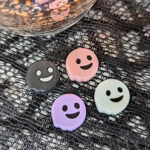 May include: Four ghost-shaped silicone phone grips in black, pink, purple, and white. Each grip has a smiling face.