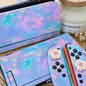 May include: A set of iridescent blue, pink, and purple Switch accessories. The set includes a console skin, a dock skin, and two controller skins. The controller skins have white flower-shaped buttons. The accessories are on a woven mat.
