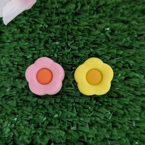 May include: Two small flower-shaped buttons, one pink with an orange center and one yellow with an orange center, on a green surface.