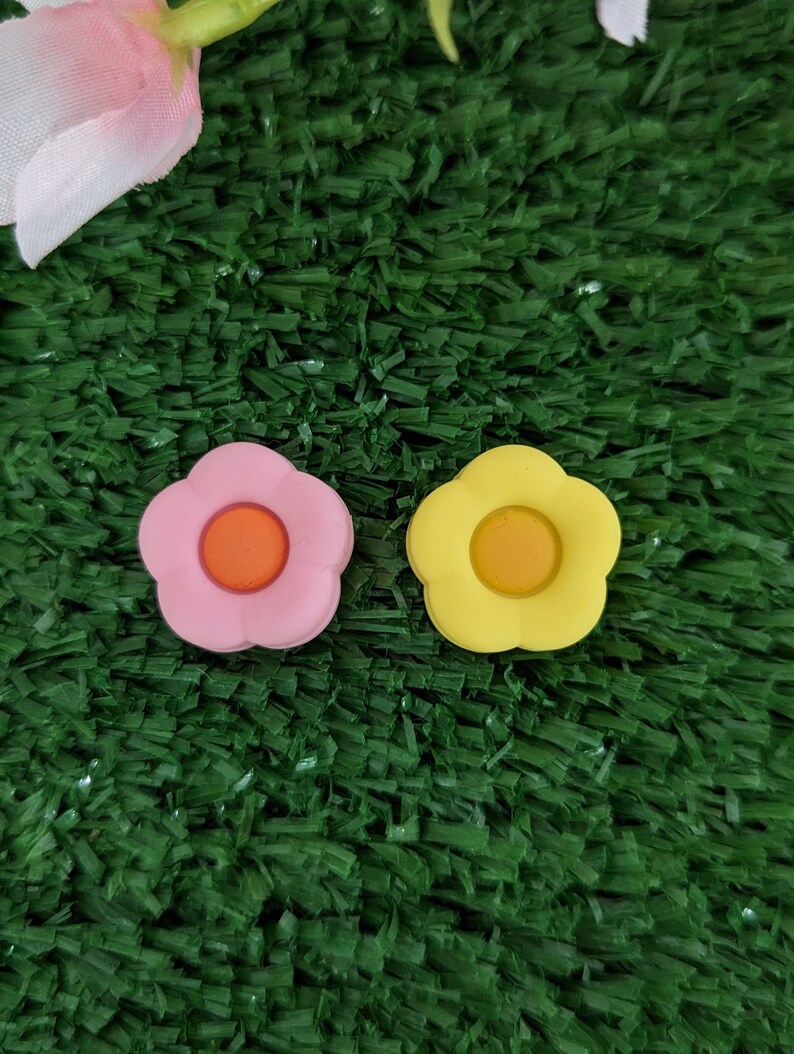 May include: Two small flower-shaped buttons, one pink with an orange center and one yellow with an orange center, on a green surface.