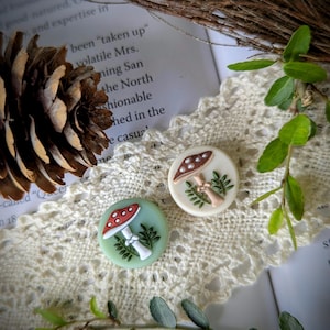 May include: Two decorative mushroom-themed buttons. One button is light green with a red and white mushroom design, and the other is off-white with a brown and white mushroom design. Both buttons have green leaf accents. The buttons are on a white lace doily.