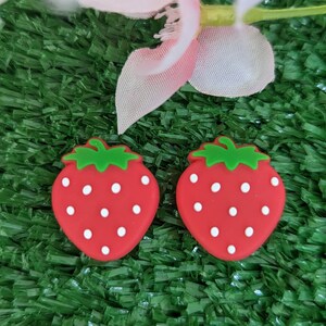 May include: Two red strawberry-shaped earrings with white dots on a green artificial grass background.