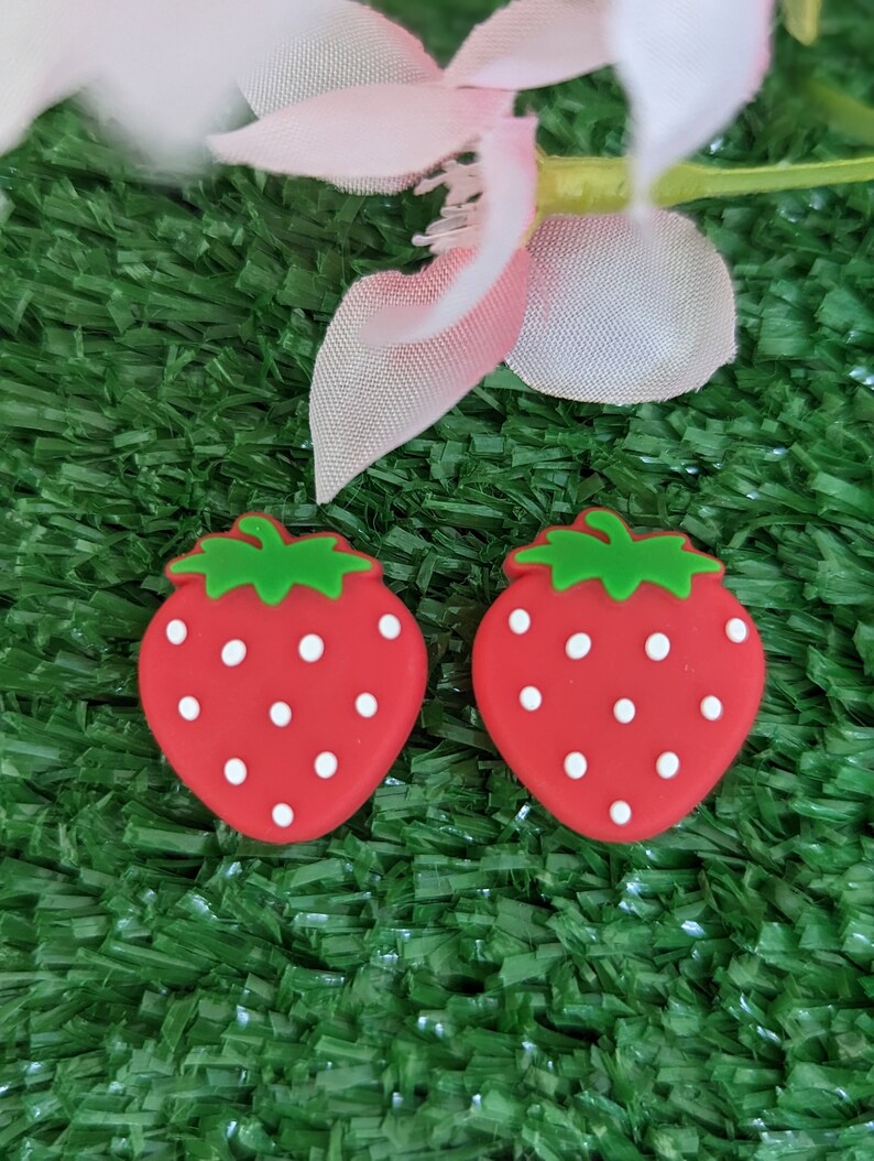 May include: Two red strawberry-shaped earrings with white dots on a green artificial grass background.