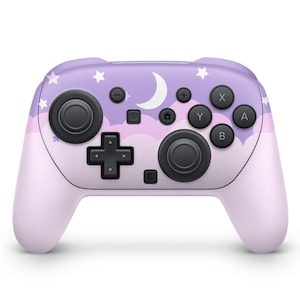 以下が含まれることがあります： ラベンダーとピンクのビデオゲームコントローラー。上部には月、星、雲の模様があり、黒いボタンとコントロールスティックが付いています。下部と側面は無地のライトピンクです。