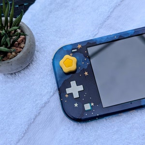May include: A blue and black Nintendo Switch Lite console with a star-themed design. The console has a yellow star-shaped button on the top left side.