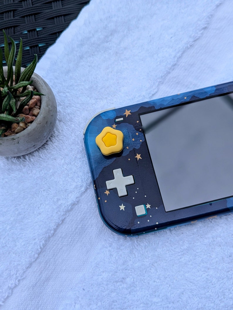May include: A blue and black Nintendo Switch Lite console with a star-themed design. The console has a yellow star-shaped button on the top left side.