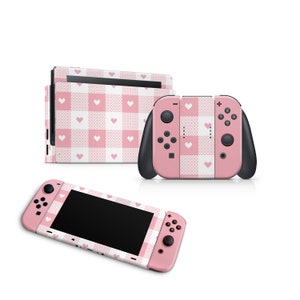 Puede incluir: Consola y mando de Nintendo Switch rosa y blanco con un patrón de cuadros y corazones blancos.