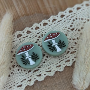 Op de afbeelding: Twee ronde, saliegroene oorbellen met een paddestoelontwerp. De paddenstoelen hebben een rode hoed met witte stippen, een witte steel en zwarte bladeren. De oorbellen liggen op een cr&egrave;mekleurig kanten lint.