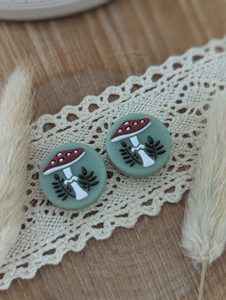 Op de afbeelding: Twee ronde, saliegroene oorbellen met een paddestoelontwerp. De paddenstoelen hebben een rode hoed met witte stippen, een witte steel en zwarte bladeren. De oorbellen liggen op een cr&egrave;mekleurig kanten lint.