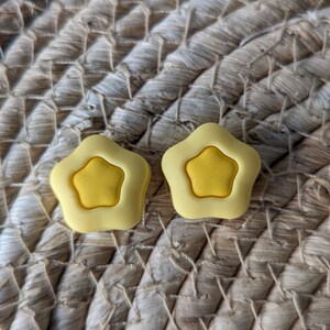 Pode incluir: Dois bot&otilde;es de silicone em forma de estrela amarela. Os bot&otilde;es est&atilde;o sobre uma superf&iacute;cie de palha tecida.