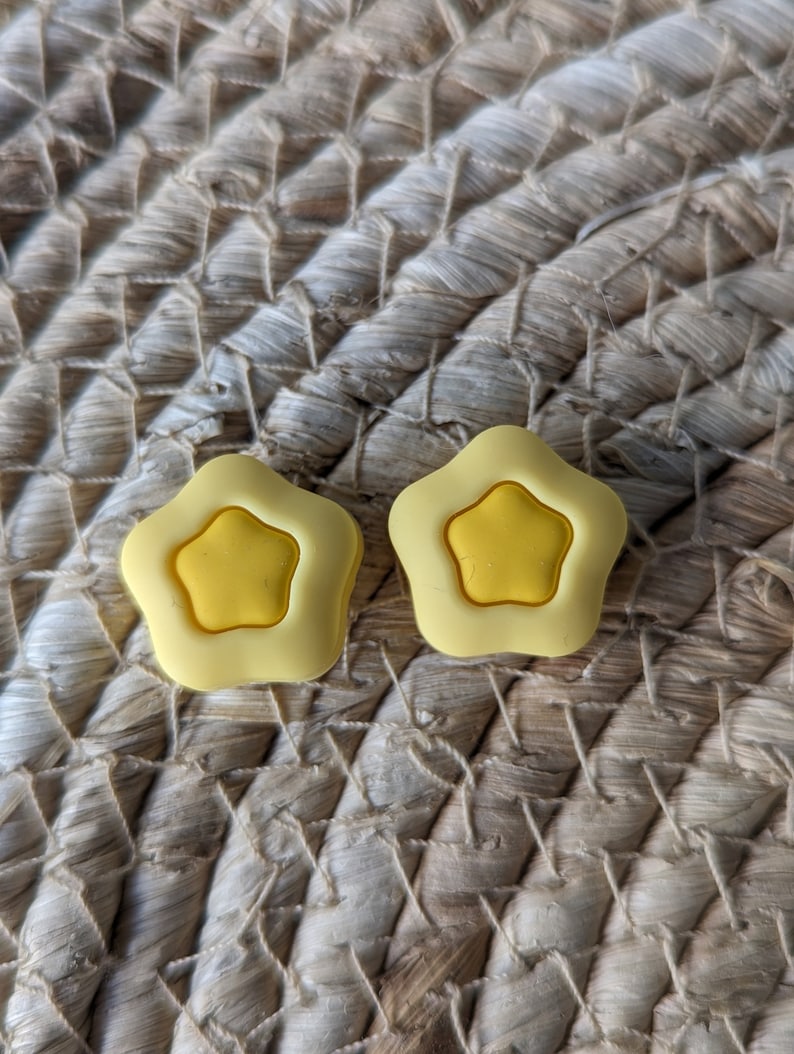 Pode incluir: Dois bot&otilde;es de silicone em forma de estrela amarela. Os bot&otilde;es est&atilde;o sobre uma superf&iacute;cie de palha tecida.