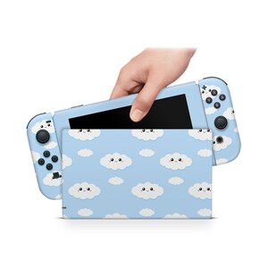 Op de afbeelding: Een blauwe en witte Nintendo Switch-consolehuid met een patroon van schattige, glimlachende wolken. De huid is ontworpen om te passen op de console en de Joy-Con-controllers.