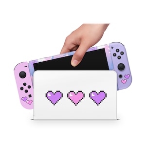 Op de afbeelding: Een roze en paarse Nintendo Switch-console met een gepixelde hartvorm. De console wordt in een witte standaard geplaatst met drie gepixelde harten aan de voorkant.