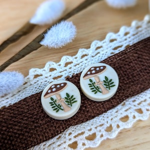 May include: A pair of white round earrings with a brown mushroom and green leaves design.