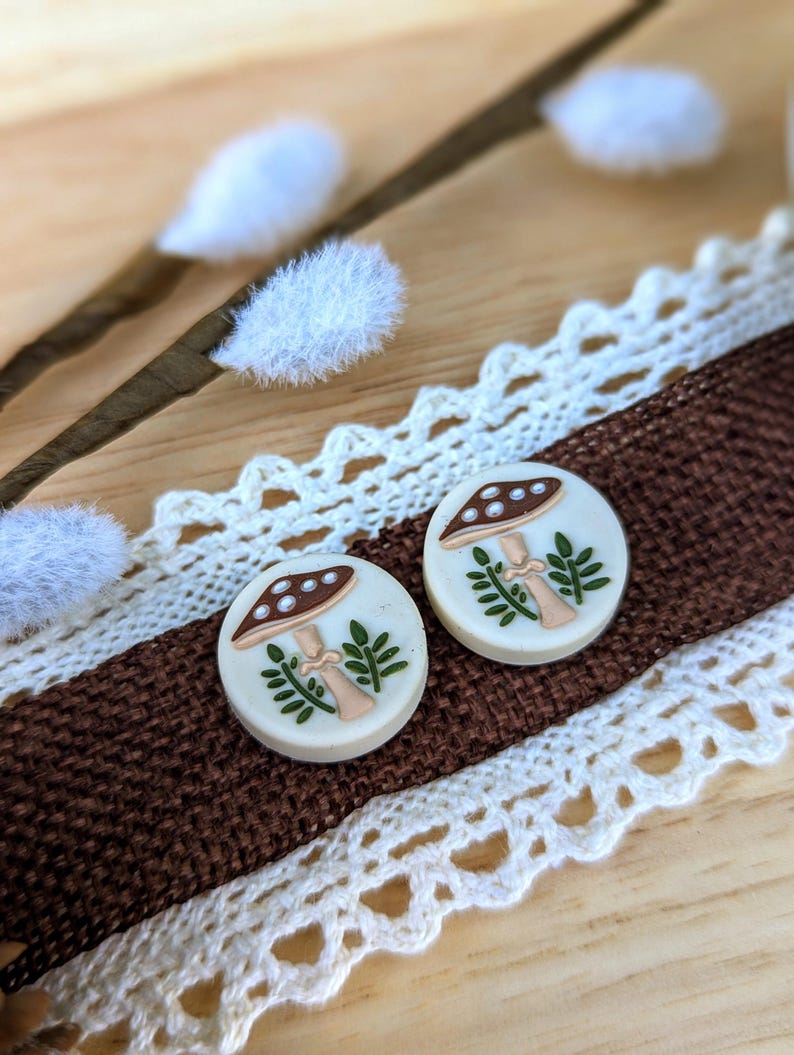 May include: A pair of white round earrings with a brown mushroom and green leaves design.