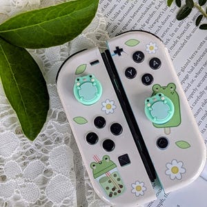 以下が含まれることがあります： 緑色のカエル、花、タピオカミルクティーが描かれたカスタムデザインの白いニンテンドースイッチ本体。本体には薄い青色のカエル型のボタンカバーが2つ付いている。