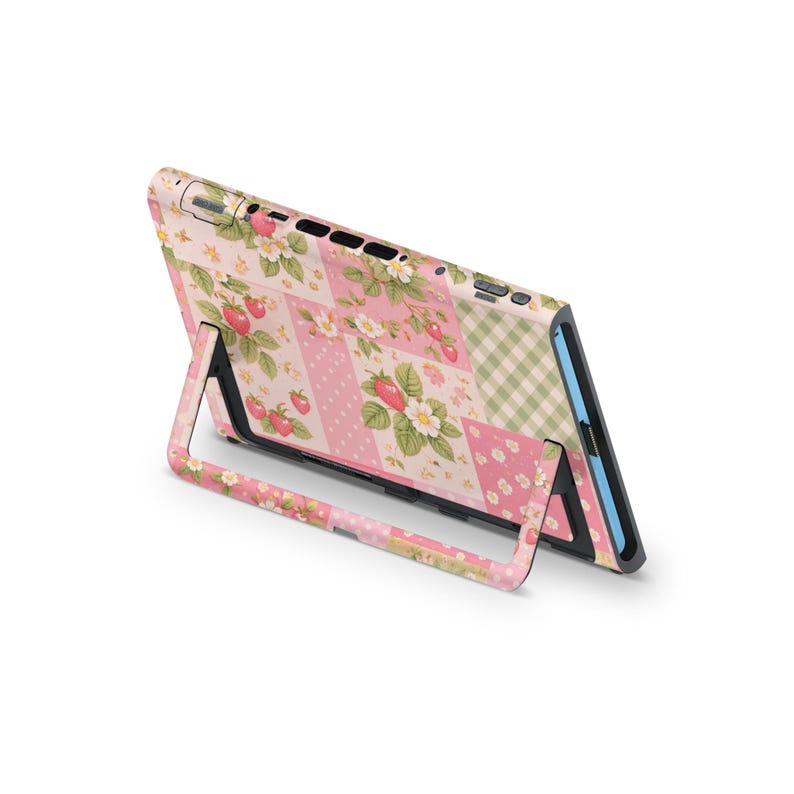 May include: A handheld gaming device case with a pink and white patchwork design featuring strawberries, flowers, and a gingham pattern. The device is supported by a matching stand.