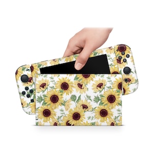 Puede incluir: Una consola Nintendo Switch blanca con una piel de patrón de girasol. La consola está cerrada y la pantalla es visible. La piel presenta girasoles amarillos con centros marrones y hojas verdes sobre un fondo blanco.