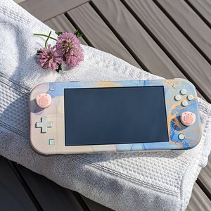 May include: A blue, white, and gold Nintendo Switch Lite console with pink buttons. The console is lying on a white towel.