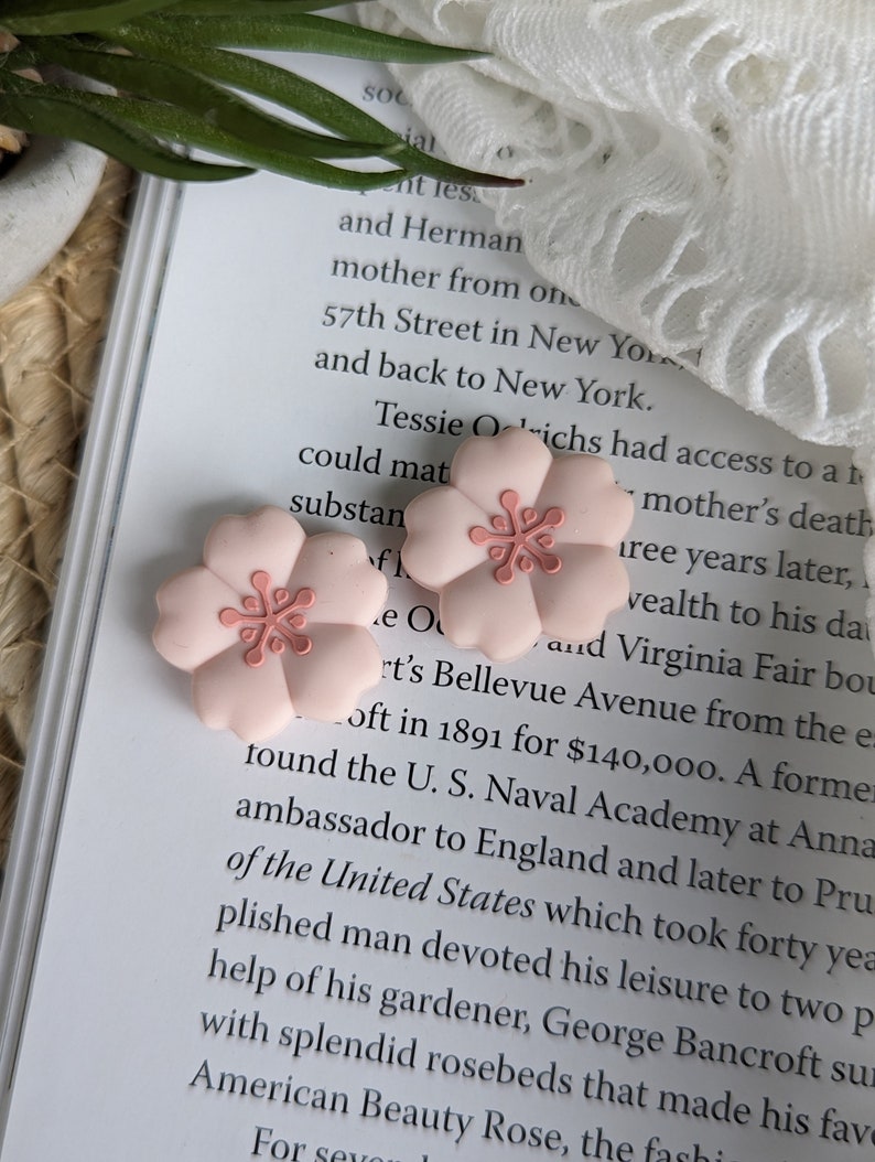 May include: Two pink flower-shaped hair clips with a pink flower design in the center. The hair clips are on a white surface with text in the background.