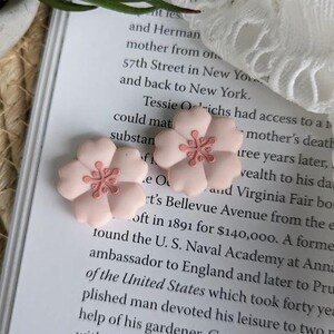 May include: Two pink ceramic flower hair clips with a light pink center. The flowers have a 3D design and are perfect for adding a touch of whimsy to any hairstyle.