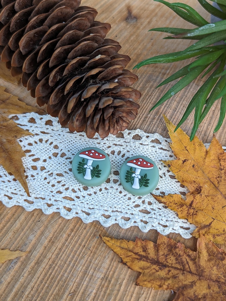 Op de afbeelding: Twee ronde oorbellen met een paddenstoelontwerp. De oorbellen hebben een lichtgroene achtergrond met een rood-witte paddenstoelillustratie. De oorbellen liggen op een wit kanten kleedje, met een dennenappel en herfstbladeren op de achtergrond.