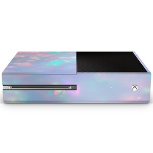 Puede incluir: Una consola Xbox One blanca con un dise&ntilde;o de &oacute;palo iridiscente. La consola tiene un panel frontal negro con un dise&ntilde;o texturizado.