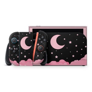 以下が含まれることがあります： 黒とピンクのNintendo Switchコンソール。黒い背景にピンクの月、星、雲が描かれたデザインです。コントローラーも同じデザインです。白い背景に置かれています。