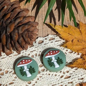 Op de afbeelding: Twee ronde, groene oorbellen met rode en witte paddenstoelmotieven. Ze liggen op een achtergrond van een dennenappel, kant en herfstbladeren. Een accessoire met een natuurthema.