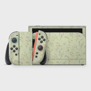 Peut inclure: Une console Nintendo Switch et ses manettes avec un motif floral. Les manettes sont détachées, révélant les boutons et les joysticks. Le motif floral présente de petites fleurs roses, jaunes et blanches sur un fond vert clair.
