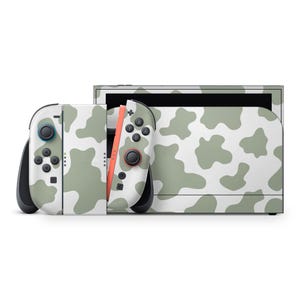 Może przedstawiać: Biała konsola Nintendo Switch i kontrolery z zielonym wzorem w krowie łaty. Kontrolery mają czarne i czerwone akcenty. Konsola znajduje się na białym tle.