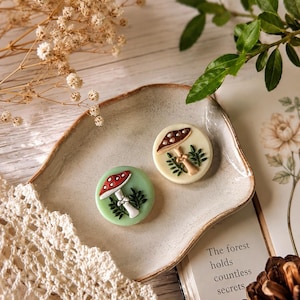 May include: Two circular buttons featuring raised mushroom designs. One button is light green with a red-capped mushroom, while the other is cream-coloured with a brown-capped mushroom. Both display white stems and small green leaf accents.