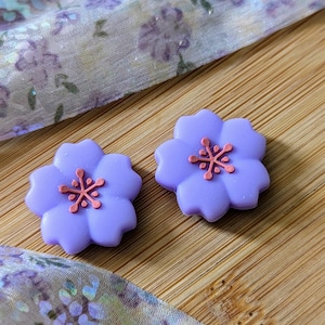 Può includere: Due abbellimenti a forma di fiore color lavanda con centri rosa. Gli accenti floreali sono posti su una superficie di legno, con un tessuto fantasia sullo sfondo. Questi elementi decorativi potrebbero essere utilizzati per l'artigianato o la gioielleria.