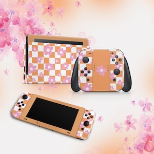 Può includere: Un set di console Nintendo Switch e controller Joy-Con a scacchi arancioni e bianchi con accenti di fiori rosa.