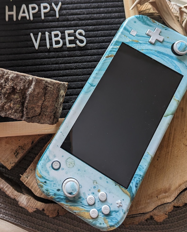 May include: A blue and gold Nintendo Switch Lite with a marble design. The console is sitting on a wooden surface with a black letter board that says "HAPPY VIBES".