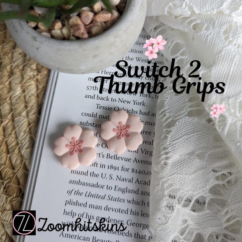Puede incluir: Dos agarres para el pulgar en forma de flor rosa para un controlador Switch 2, con un centro rosa m&aacute;s oscuro. Los agarres se muestran en un libro con texto y un fondo de encaje blanco. El texto "Switch 2 Thumb Grips" es visible.