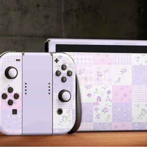 Puede incluir: Una consola Nintendo Switch y mandos con un diseño de patchwork floral en lavanda y blanco. Los mandos tienen botones y joysticks negros. La consola está sobre una superficie de madera. El diseño presenta detalles florales.